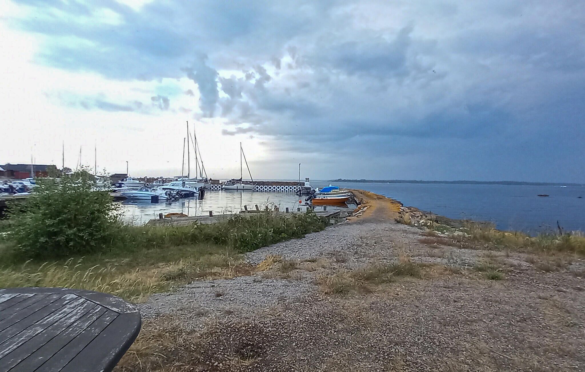 Un port de plaisance avec des bateaux amarrés, une passerelle en bois menant à l’eau et une vue lointaine sur la mer sous un ciel nuageux.