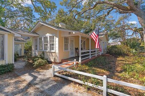 A yellow house with a porch, a white fence, and an American flag. - Beach Cottage Steps to Sand | Walk to Pier & King and Prince (Saint Simons Island)