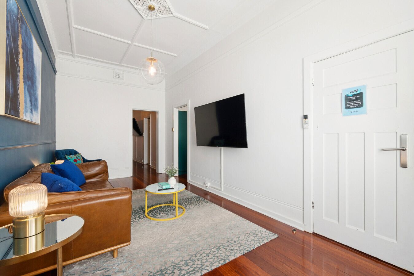 Un salon moderne avec un canapé en cuir marron, une petite table basse ronde, un téléviseur à écran plat fixé au mur et une porte blanche avec une pancarte.