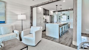 Una sala moderna con sofá, sillón y mesa de centro. Se ve una cocina al fondo, con armarios blancos y una barra.