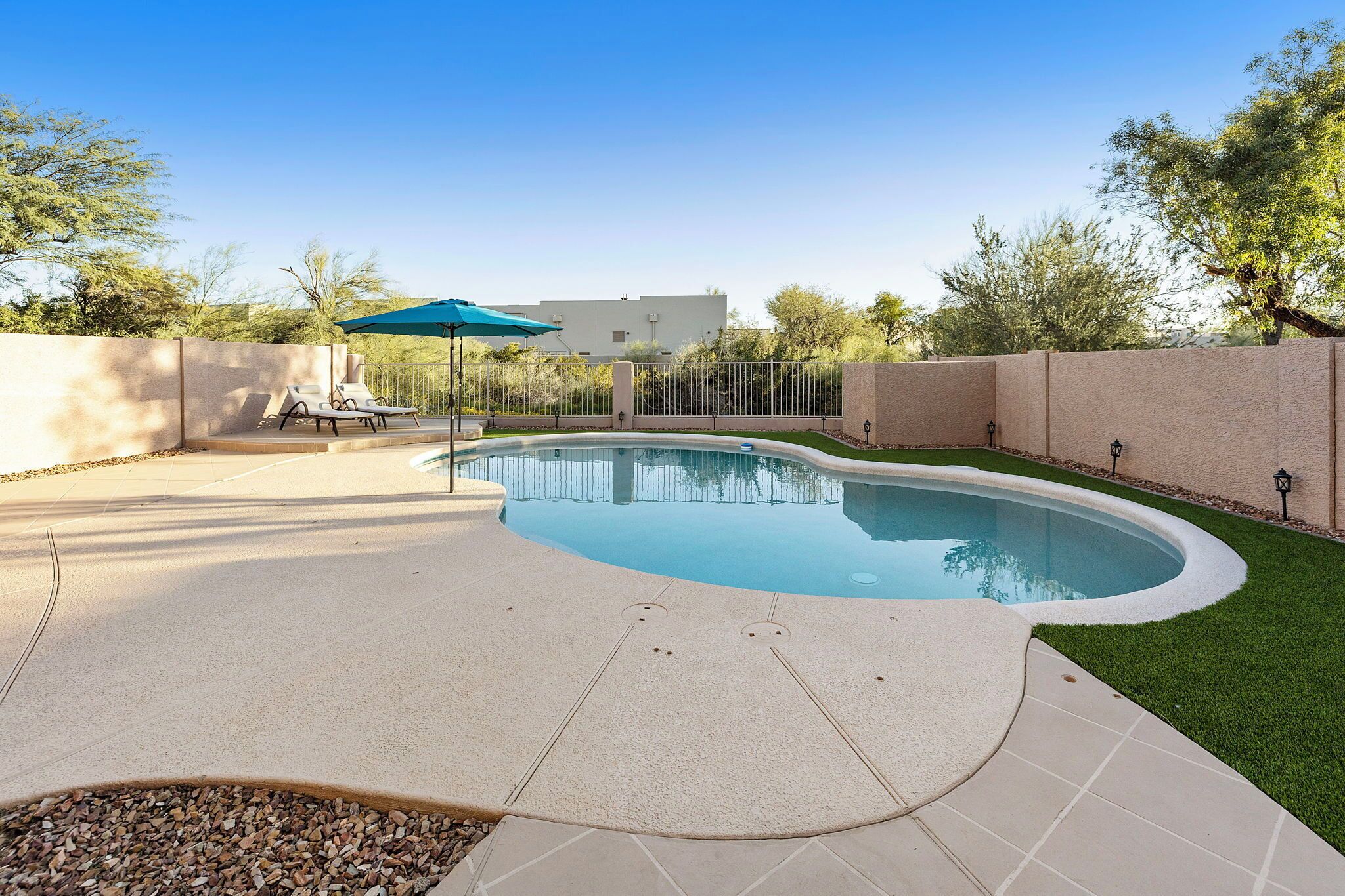 Ein Pool mit bepflastertem Rand, eine Lounge-Bereich mit Stühlen und Sonnenschirm, und eine Zähe umgibt den Bereich.
