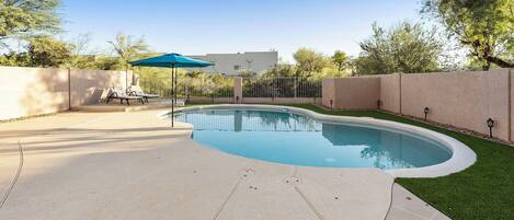 Ein Pool mit bepflastertem Rand, eine Lounge-Bereich mit Stühlen und Sonnenschirm, und eine Zähe umgibt den Bereich.
