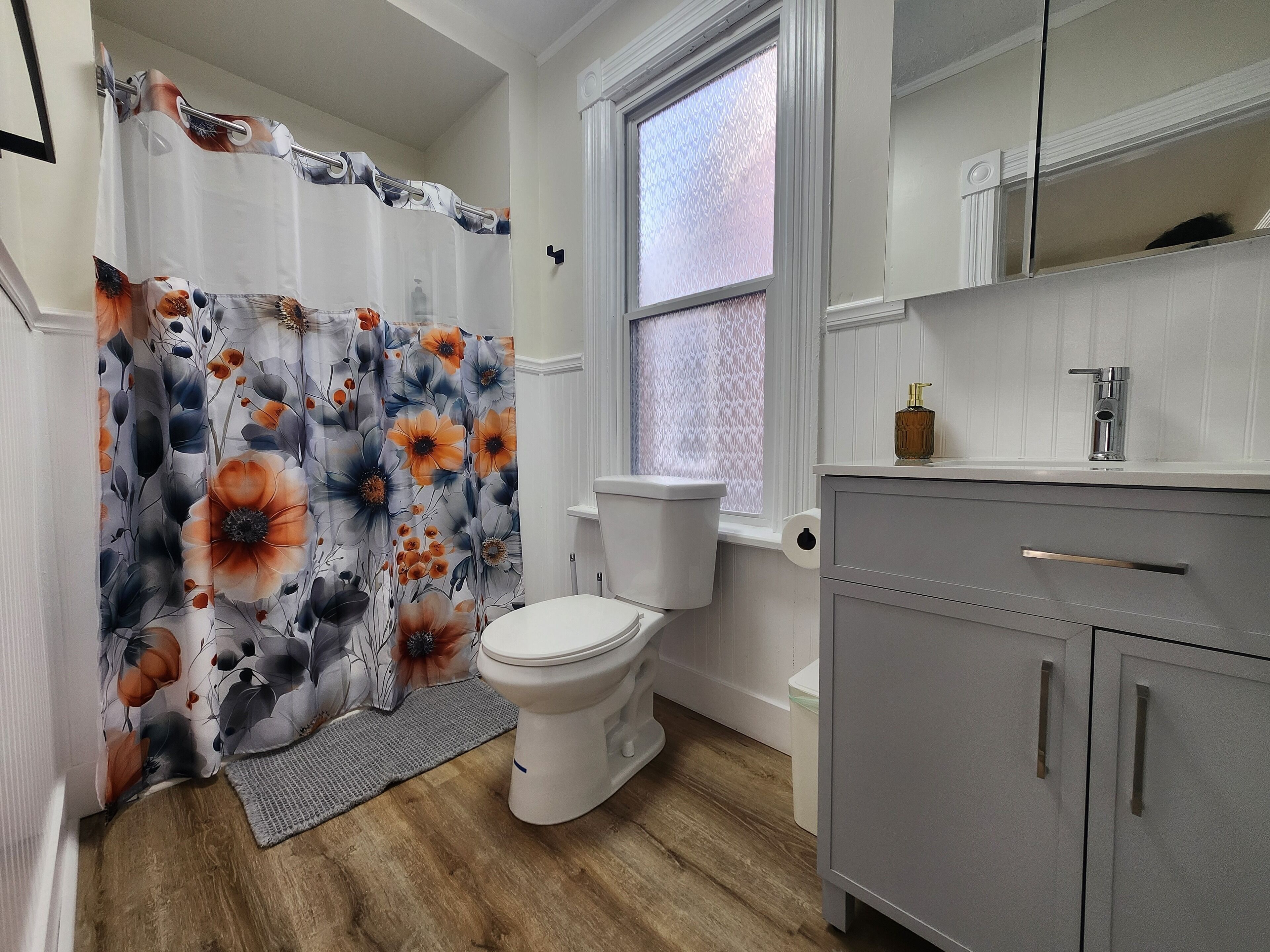 A bathroom with a toilet, a grey cabinet, a shower curtain with a floral pattern, and a window with frosted glass.