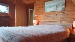 Een slaapkamer in een houten hutje, met een bed, nachtkastje, lamp en een schilderij van een strand.