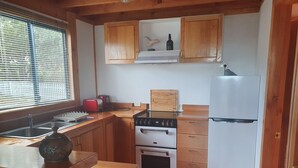 Een compacte keuken met houten kastjes, een inbouwoven, een koelkast en een raam met zonwering.