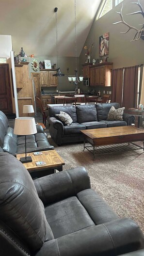 A living room with a sofa, coffee table, and lamp. There is a kitchen in the background with cabinets and a dining area. - Whispering Pines Cabin, Hot Tub, Beach Access, Game Room, Sleeps 8 (McCall)