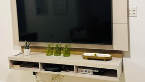 A flat-screen TV mounted on a white wall with a wooden panel above it. Below the TV, there are three green glass bottles, a small potted plant, and a clock.