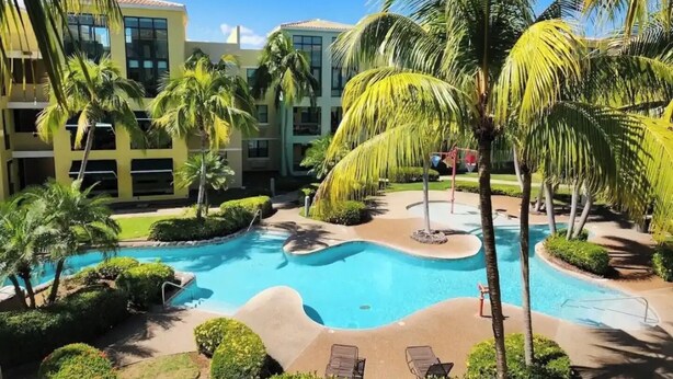 A resort with a central swimming pool, palm trees, and a building with multiple balconies.