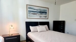 A bedroom with a bed, a nightstand with a lamp, a dresser, and a framed picture on the wall.