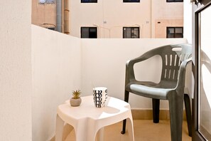 Un pequeño balcón con una mesa blanca, una planta en maceta, una taza negra y blanca y una silla de plástico gris.