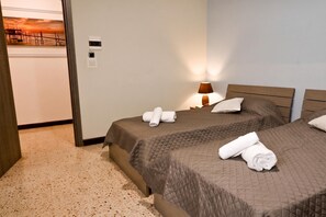 Habitación de hotel con dos camas, una mesita de noche con lámpara, un termostato en la pared y una puerta que da a otra habitación.