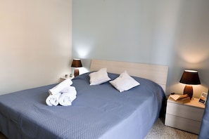 A neatly made bed with pillows, a bedside lamp, a small table with a book and a picture, and a wall-mounted clock.