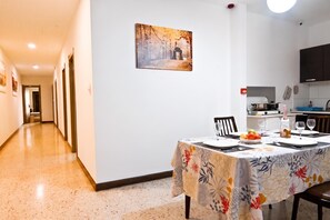 A dining area with a table set for two, a painting on the wall, and a kitchen in the background. - Sunny Ensuite by the Sea - A5, Saint Paul's Bay, Malta (Saint Paul's Bay)