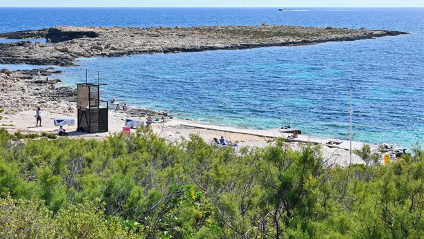 Ein Küstenstrand mit Felsformationen, klarem blauen Wasser und spärlicher Vegetation.