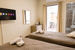A hotel room with two beds, a TV, a mirror, a small table with a vase, and a view of the outside. - Sunny Ensuite by the Sea - A4, Saint Paul's Bay, Malta (Saint Paul's Bay)