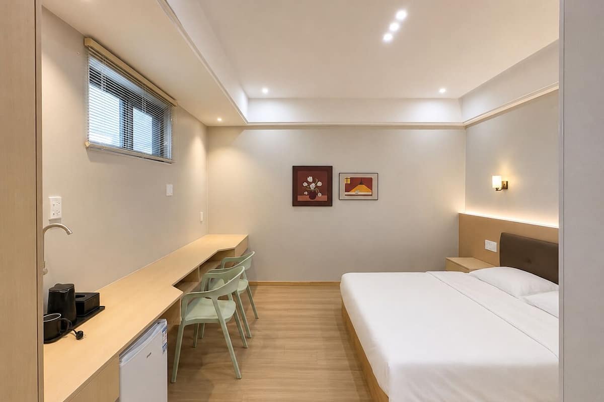 A compact hotel room with a bed, a desk with chairs, a small kitchenette, and a window with blinds.