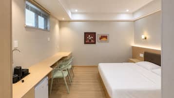 A compact hotel room with a bed, a desk with chairs, a small kitchenette, and a window with blinds.