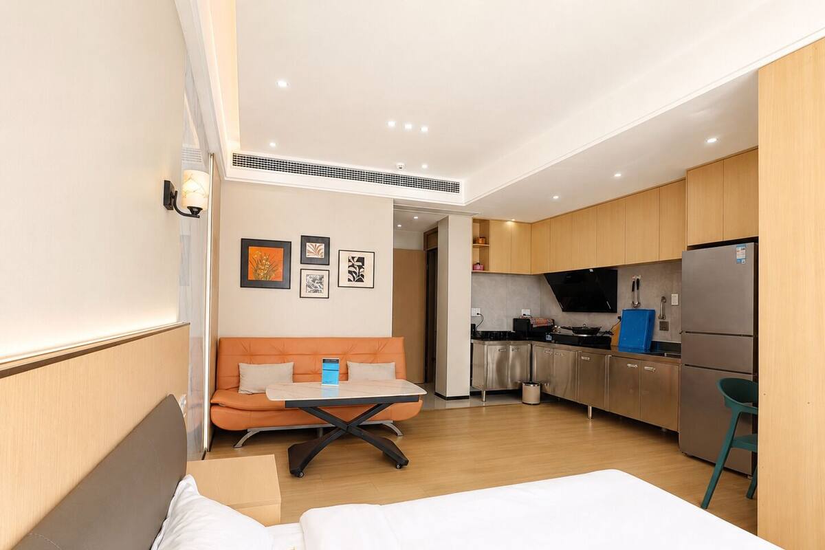 A compact hotel room with a bed, a small table, a sofa, a kitchenette, and a bathroom visible through an open door.