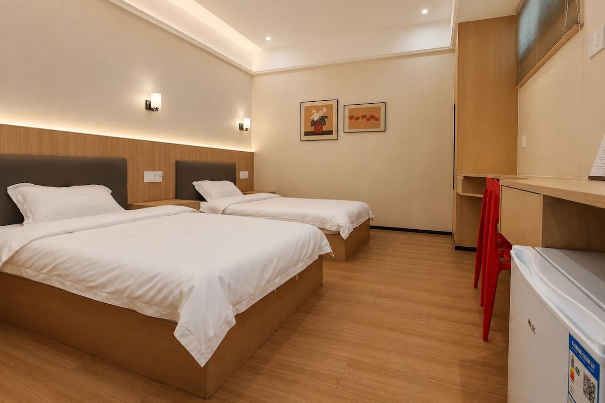 A hotel room with two beds, a desk, a refrigerator, and artwork on the wall.