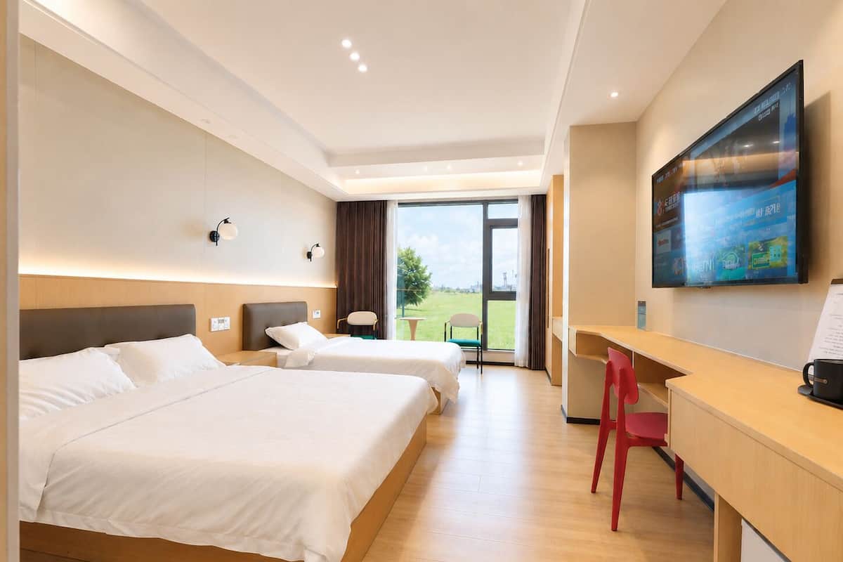 A hotel room with two beds, a desk, a chair, and a large window offering a view of the outdoors.