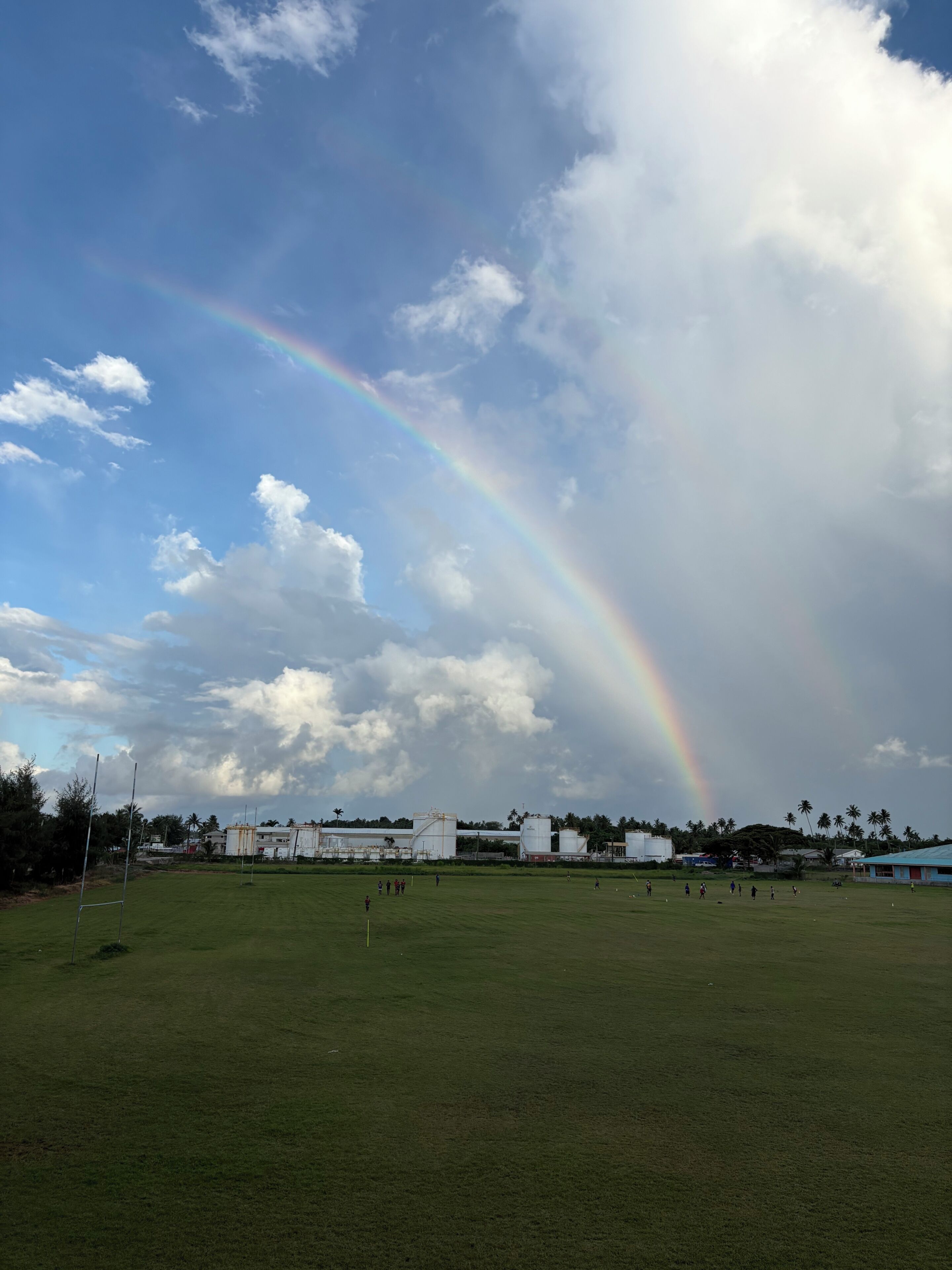 A grassy field with a rainbow, a few buildings, and a clear sky.