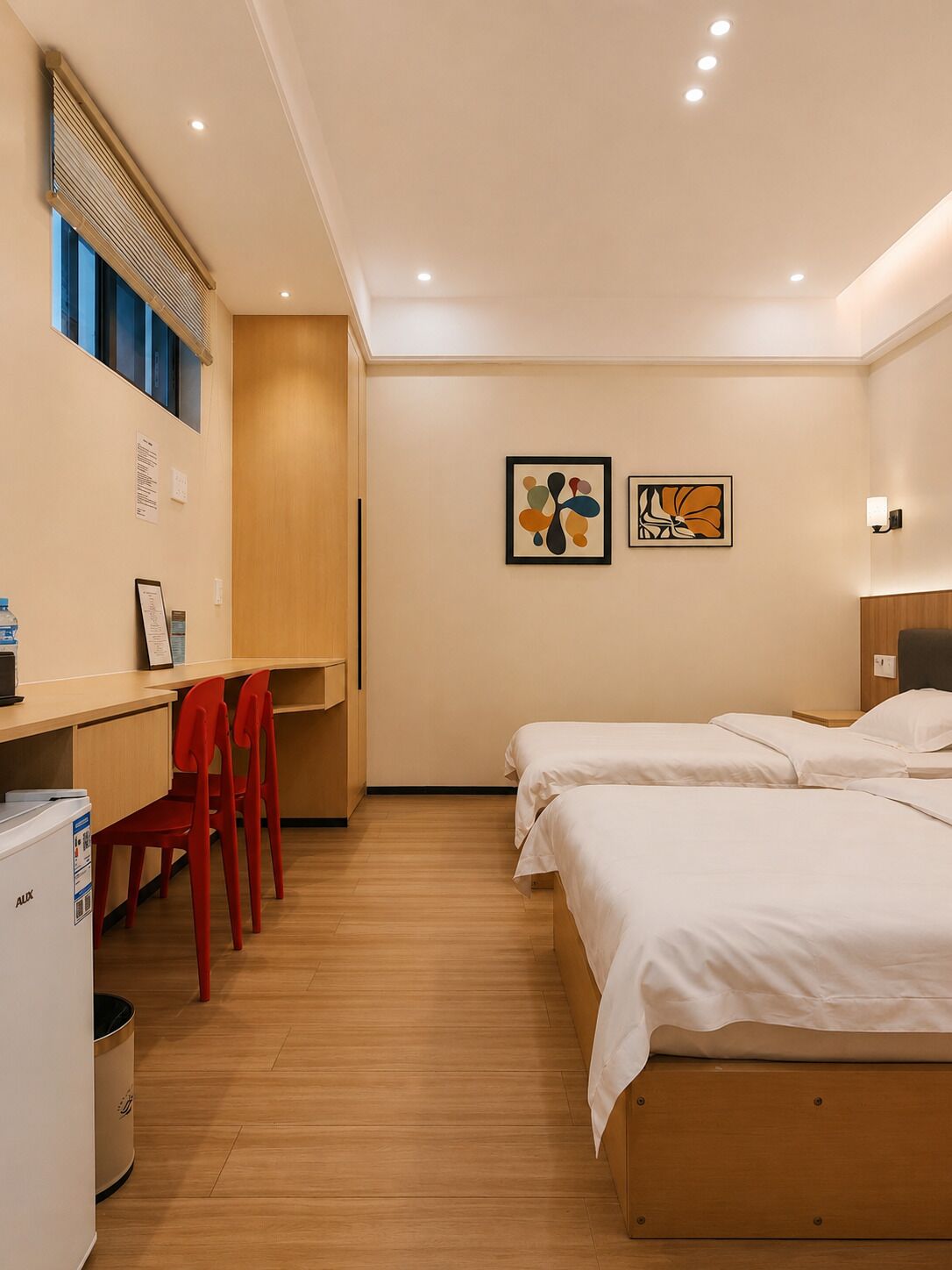 Habitación de hotel con un escritorio de madera, dos camas, una silla roja y dos obras de arte colgadas en la pared.