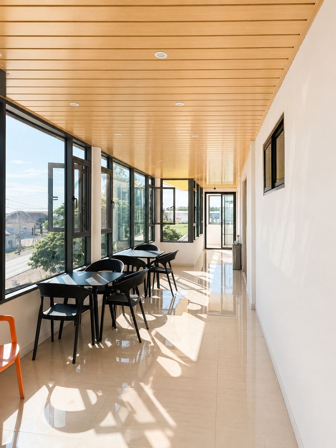 Un intérieur moderne avec un long couloir, de grandes fenêtres et un coin repas comprenant des tables et des chaises.