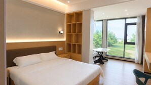 A modern hotel room with a large bed, built-in shelving, a desk, and a view of greenery through large windows. - Fantasy Inn (Nuku'alofa)