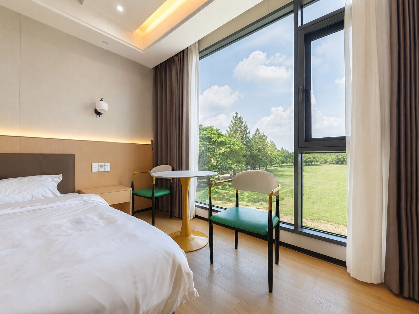 A hotel room with a large bed, a small table, two chairs, and a view of a green landscape through large windows.