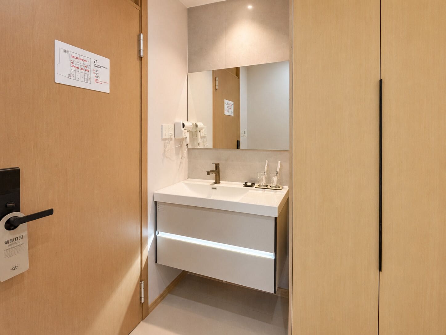 Habitación de hotel con puerta de madera, lavamanos blanco y espejo encima, además de un cartel en la pared.