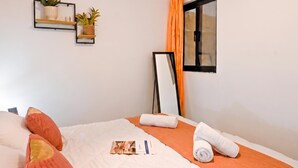 Un lit bien fait, recouvert d’une couverture blanche et orange, avec deux serviettes blanches roulées et un petit livret posé sur le lit.