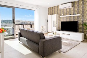 Ein modernes Wohnzimmer mit grauem Sofa, einem Flachbildfernseher und einem Balkon mit Blick auf die Stadt.
