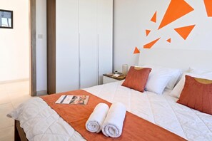 Quarto de hotel com cama, almofadas, mesa de cabeceira, revista e toalhas dobradas.