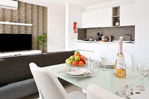 Uma sala de estar moderna com sofá, mesa de jantar de vidro para dois e TV na parede.