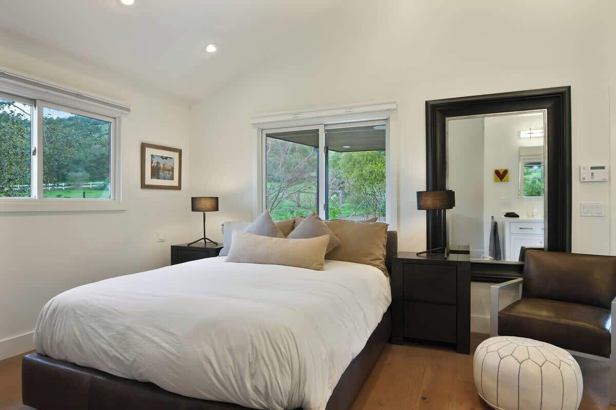 Un dormitorio con una cama grande, dos mesitas de noche, una silla, un espejo y ventanas con vistas al follaje.