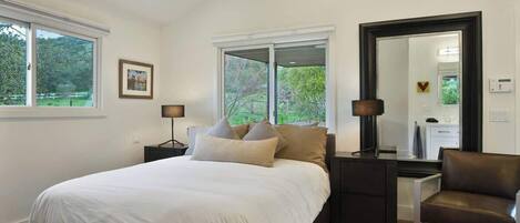 Un dormitorio con una cama grande, dos mesitas de noche, una silla, un espejo y ventanas con vistas al follaje.