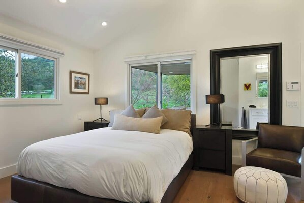 Une chambre à coucher avec un grand lit, deux tables de chevet, une chaise, un miroir et des fenêtres donnant sur un paysage verdoyant.