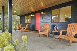 Une véranda couverte avec des chaises en bois et une petite table, une porte rouge et de grandes fenêtres.