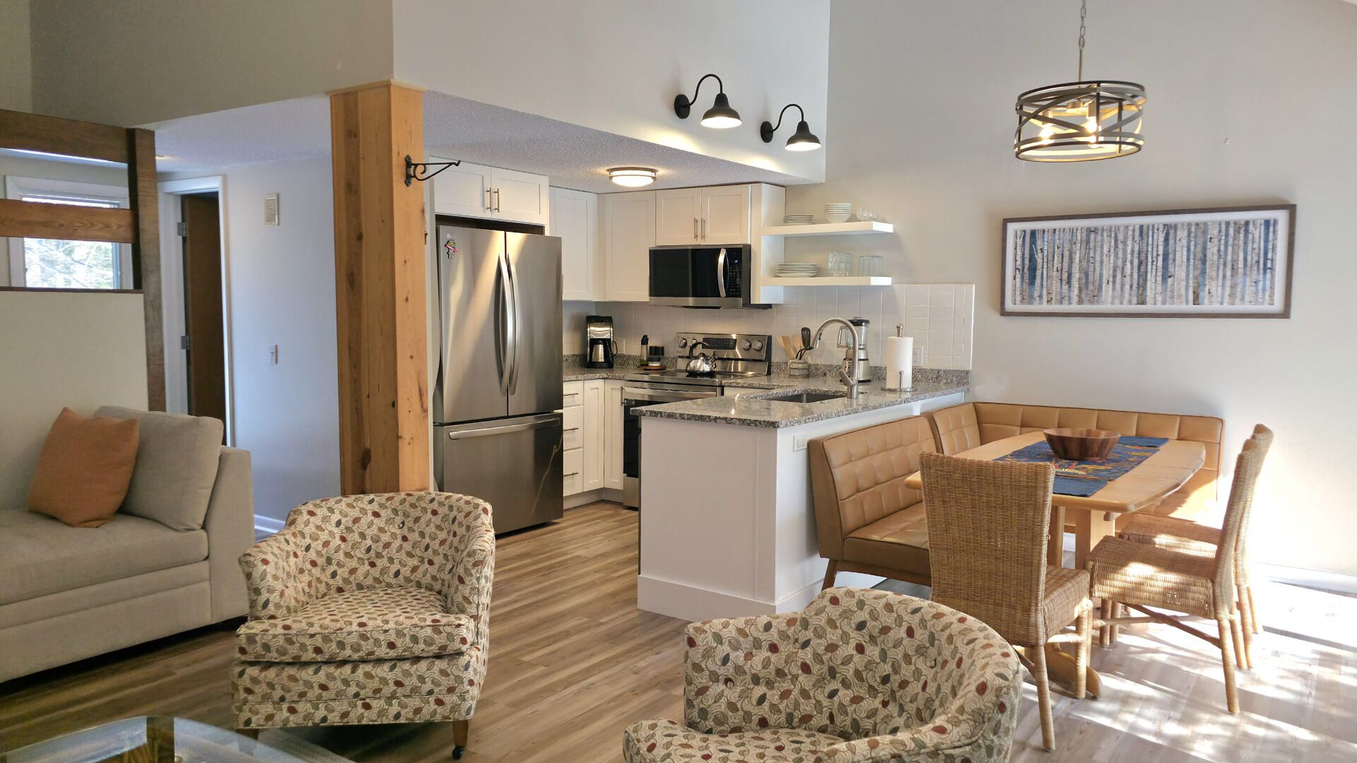 A modern kitchen with white cabinets, stainless steel appliances, and a central island. There is a dining area with a table and chairs, and a living room with a sofa and patterned chairs.
