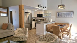 Eine moderne Küche mit weißen Schränken, Edelstahlgeräten und einer zentralen Kochinsel. Es gibt einen Essbereich mit Tisch und Stühlen sowie ein Wohnzimmer mit einer Couch und gemusterten Sesseln.