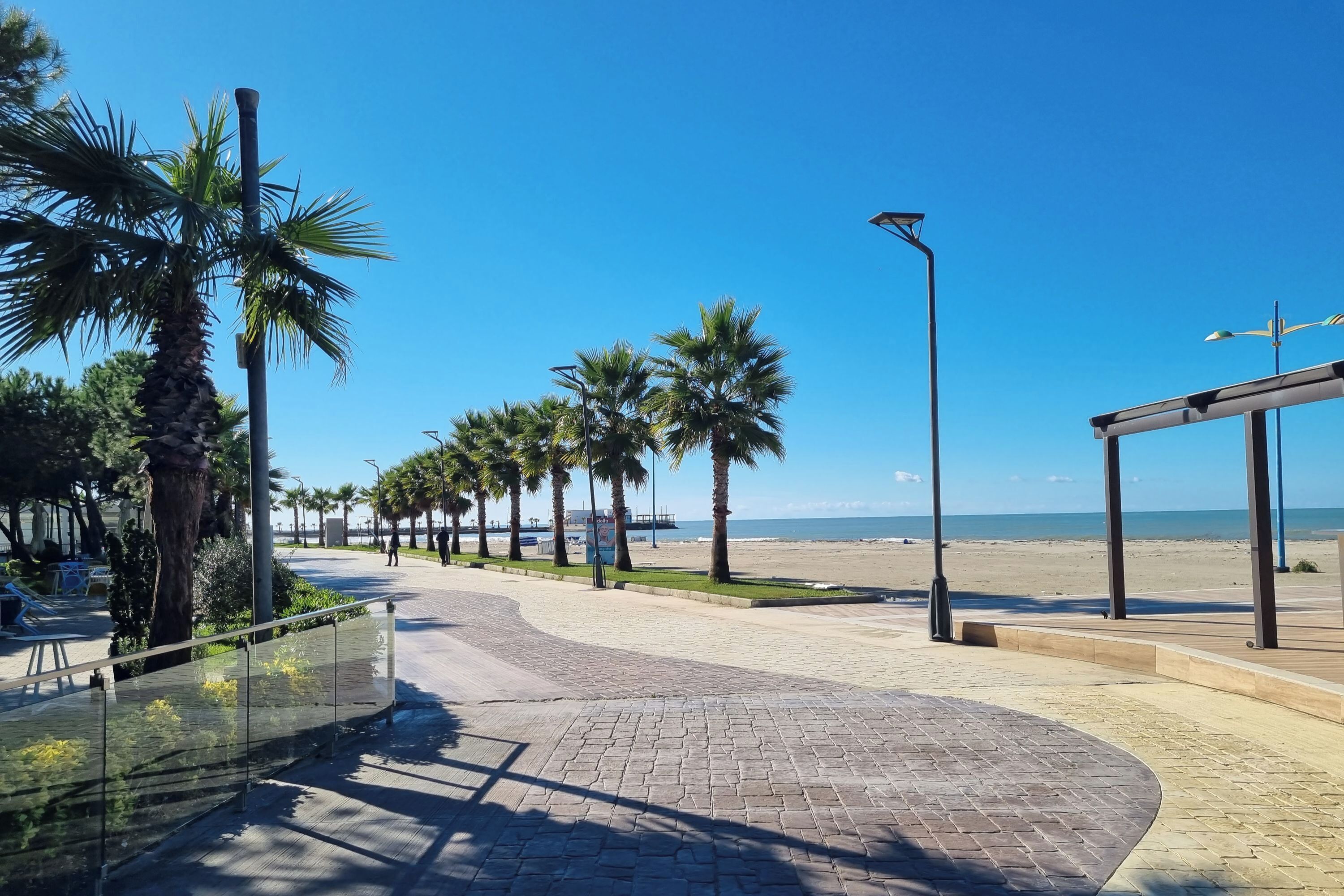Une promenade dallée bordée de palmiers, une plage en arrière-plan et un ciel bleu azur.