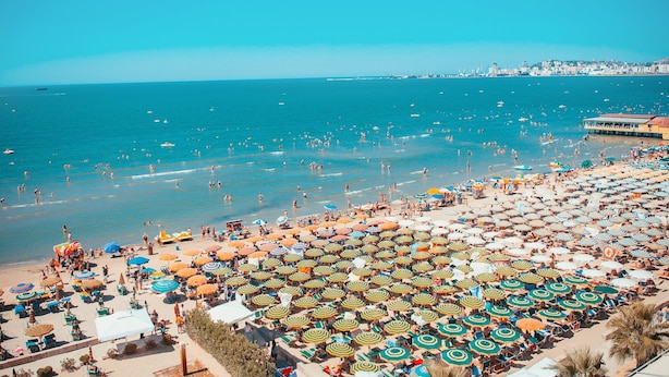 A beach with numerous umbrellas and sun loungers, a clear blue sea, and a distant cityscape.