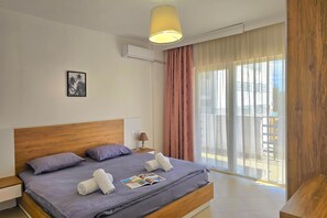 A modern hotel room with a wooden headboard, a bed with grey bedding, a bedside table with a lamp, and a large window with sheer curtains.