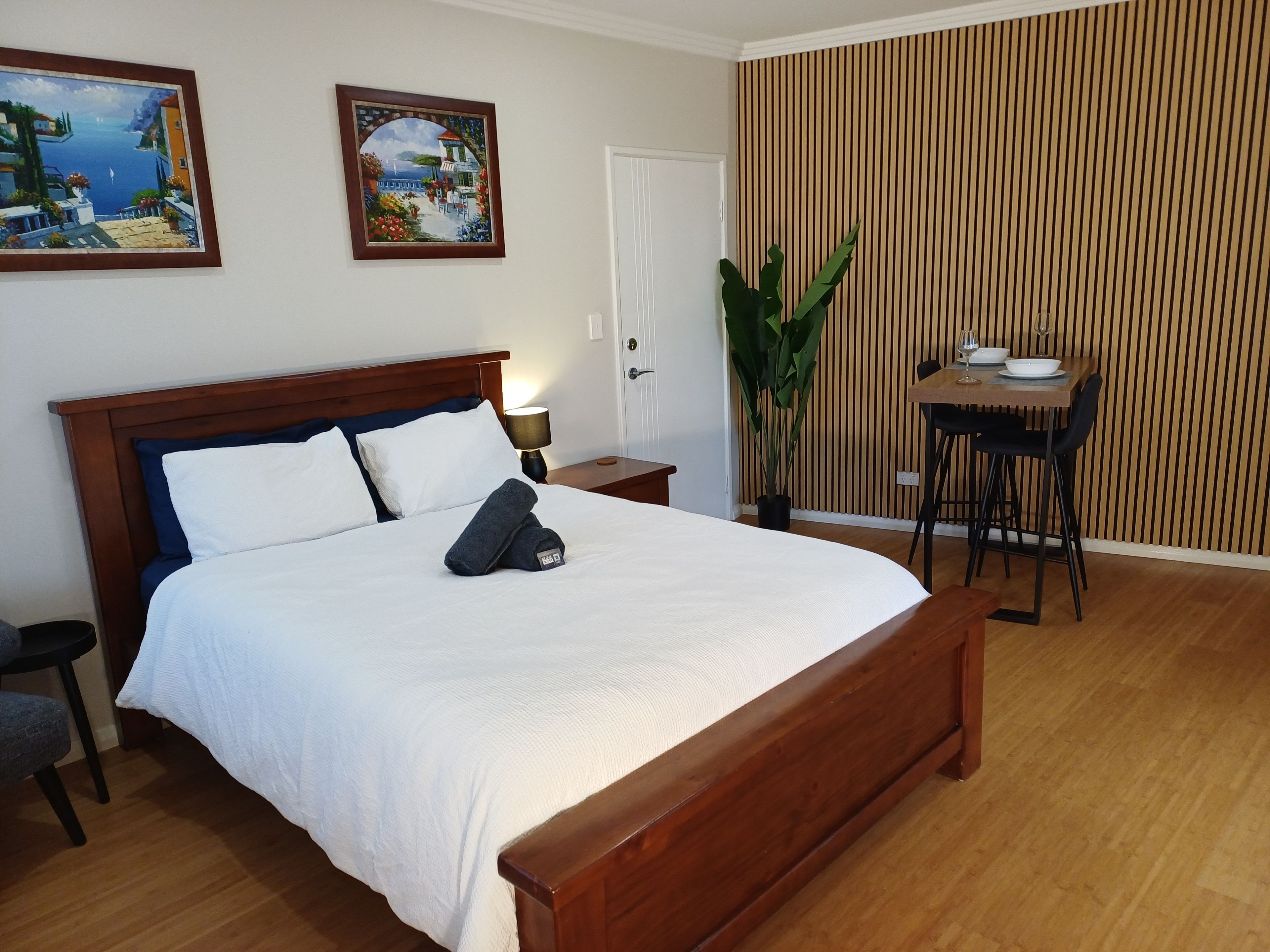 A hotel room with a large bed, a desk, and a chair. There is a painting on the wall and a potted plant.