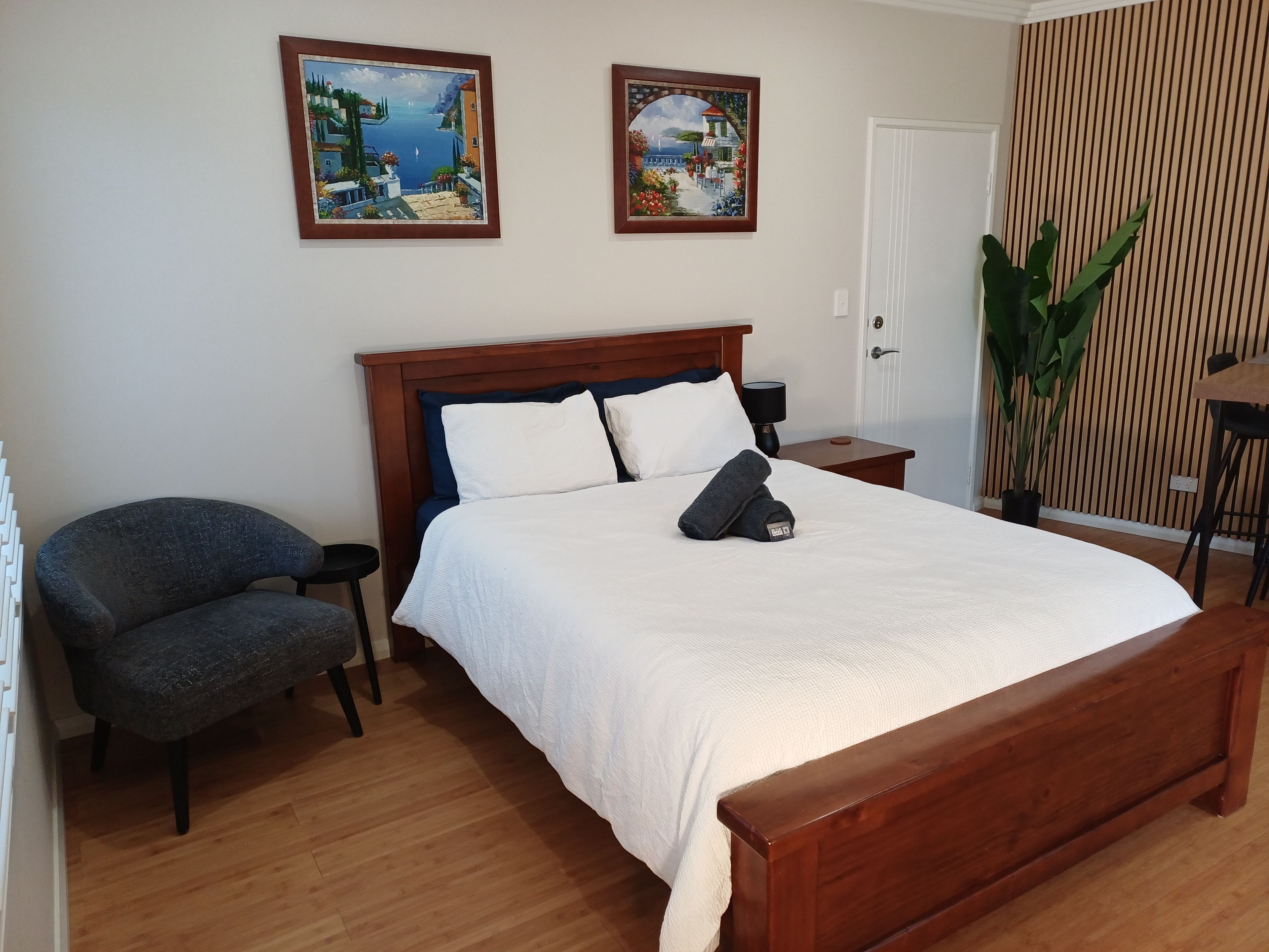 A neatly arranged bedroom with a wooden bed, a chair, a nightstand, and two wall-mounted paintings.