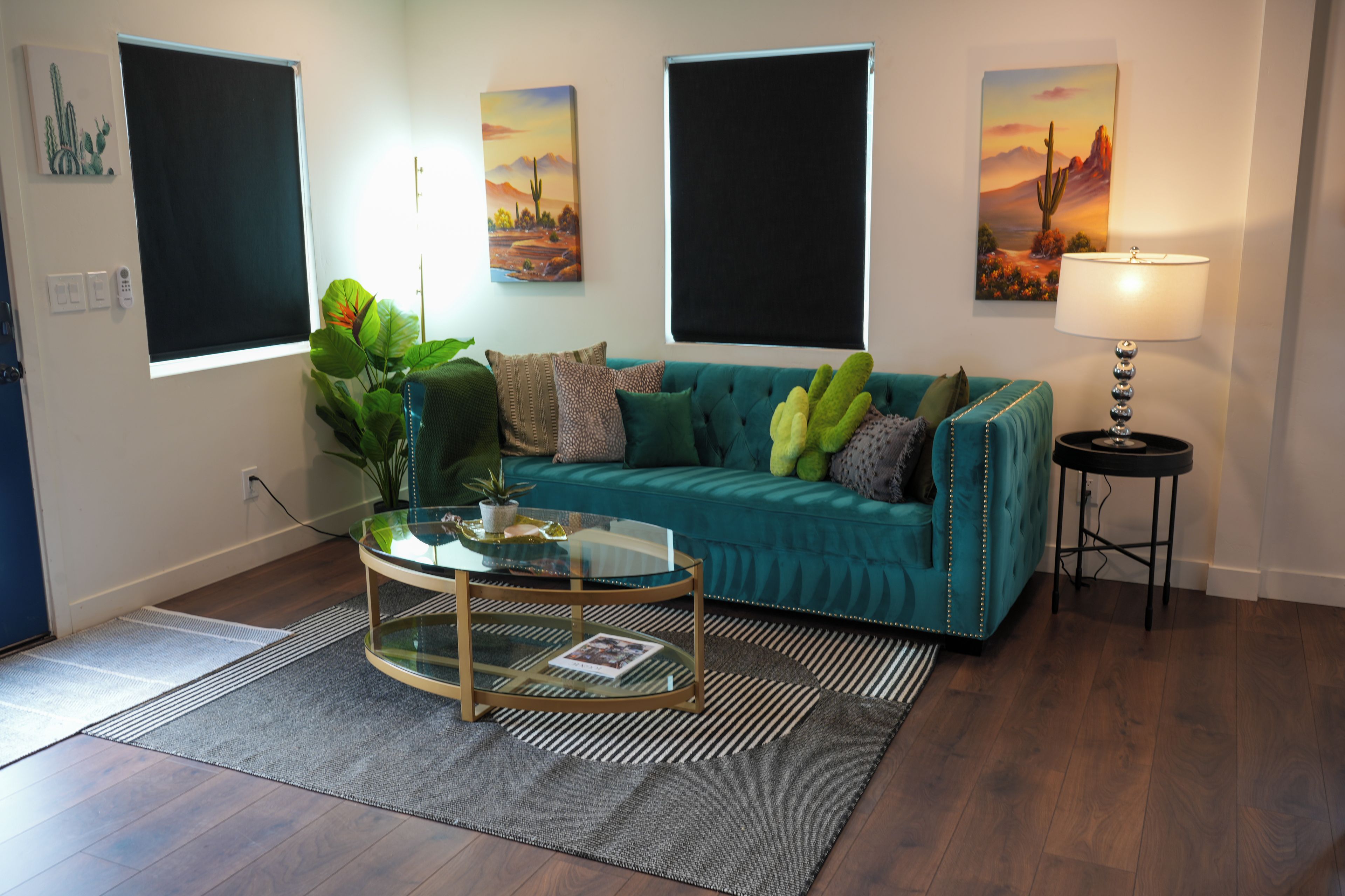 A living room with a teal sofa, a glass coffee table, and a lamp. There are paintings on the walls and a plant on the floor.