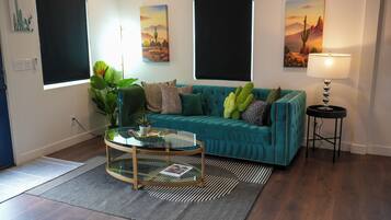 A living room with a teal sofa, a glass coffee table, and a lamp. There are paintings on the walls and a plant on the floor.