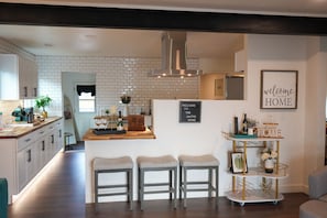 Une cuisine moderne avec un îlot central, des tabourets de bar et un panneau indiquant : « BIENVENUE CHEZ NOUS ».
