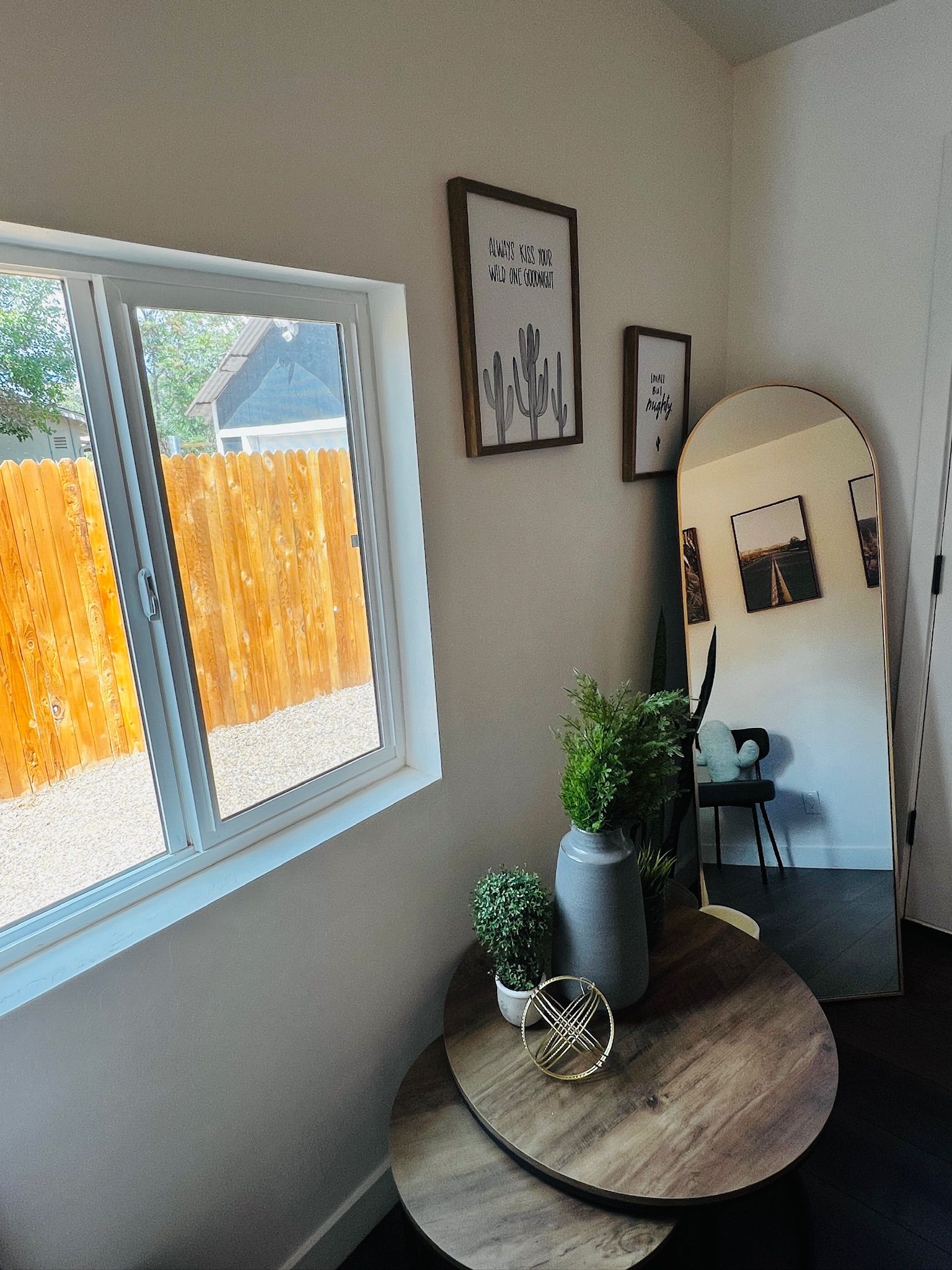 A room with a window, a round table with a potted plant, a mirror, and two framed pictures on the wall.