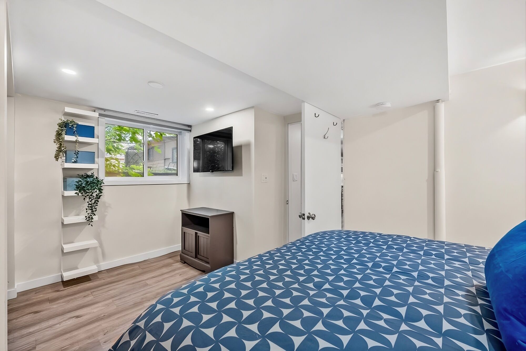 A bedroom with a bed, a TV mounted on the wall, a window with plants, and a door.